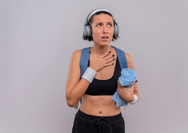 Mujer joven fitness en ropa deportiva con mochila y auriculares en la cabeza sosteniendo una toalla con la mano en el pecho mirando a un lado de pie preocupado sobre la pared blanca