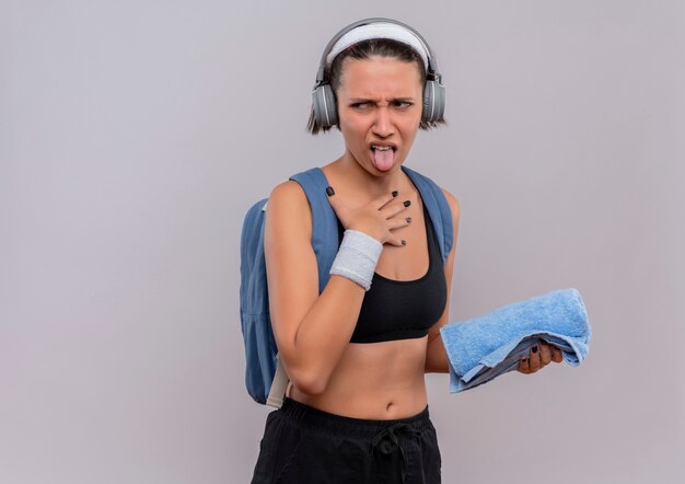 Mujer joven fitness en ropa deportiva con mochila y auriculares en la cabeza sosteniendo una toalla sacando la lengua con expresión de disgusto de pie sobre una pared blanca