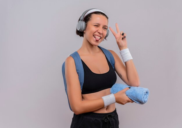 Mujer joven fitness en ropa deportiva con mochila y auriculares en la cabeza sosteniendo una toalla sacando la lengua sonriendo mostrando el signo de la victoria de pie sobre la pared blanca