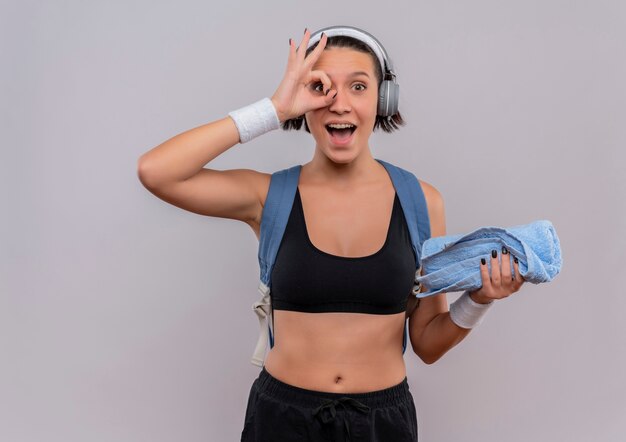 Mujer joven fitness en ropa deportiva con mochila y auriculares en la cabeza sosteniendo una toalla sonriendo alegremente