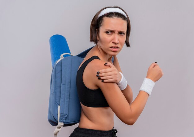 Mujer joven fitness en ropa deportiva con mochila y colchoneta de yoga que se ve mal tocando su hombro sintiendo dolor de pie sobre una pared blanca