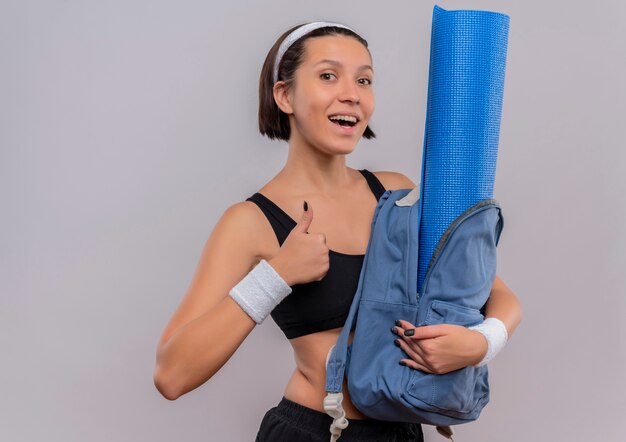 Mujer joven fitness en ropa deportiva con mochila con estera de yoga con una sonrisa en la cara mostrando los pulgares para arriba de pie sobre la pared blanca