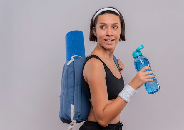 Mujer joven fitness en ropa deportiva con mochila y esterilla de yoga sosteniendo una botella de agua mirando a un lado con una sonrisa en la cara de pie sobre una pared blanca