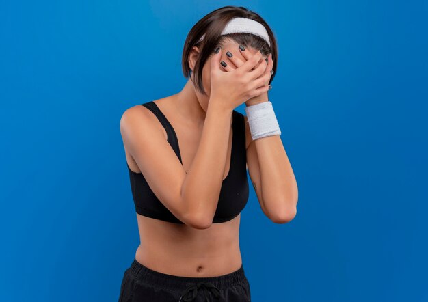 Mujer joven fitness en ropa deportiva que sufre de un fuerte dolor de cabeza que cubre la cara con las manos de pie sobre la pared azul
