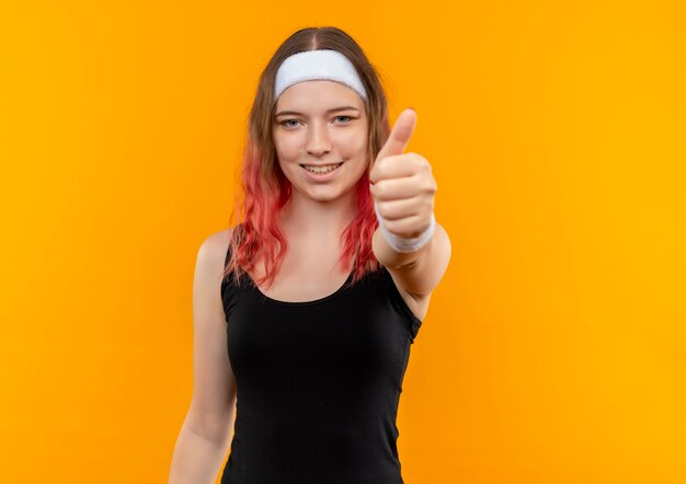 Mujer joven fitness en ropa deportiva sonriendo alegremente mostrando los pulgares para arriba de pie sobre la pared naranja