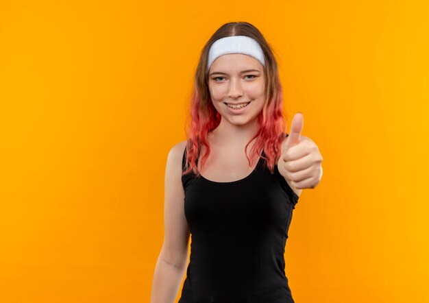 Mujer joven fitness en ropa deportiva sonriendo alegremente mostrando los pulgares para arriba de pie sobre la pared naranja