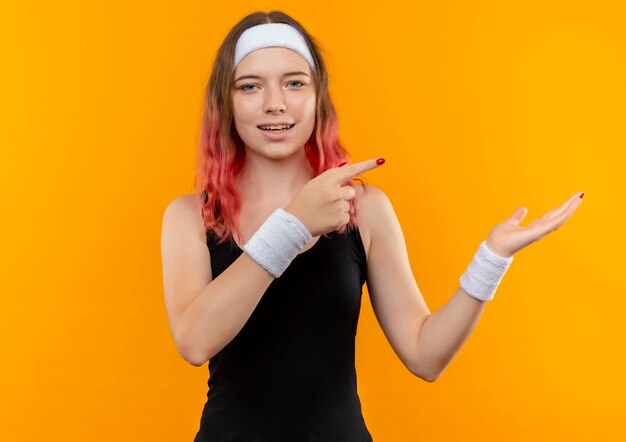 Mujer joven fitness en ropa deportiva sonriendo apuntando con el dedo índice hacia el lado que presenta con el brazo de la mano de pie sobre la pared naranja