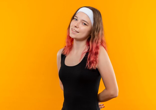 Mujer joven fitness en ropa deportiva sonriendo positivo y feliz parado sobre pared naranja