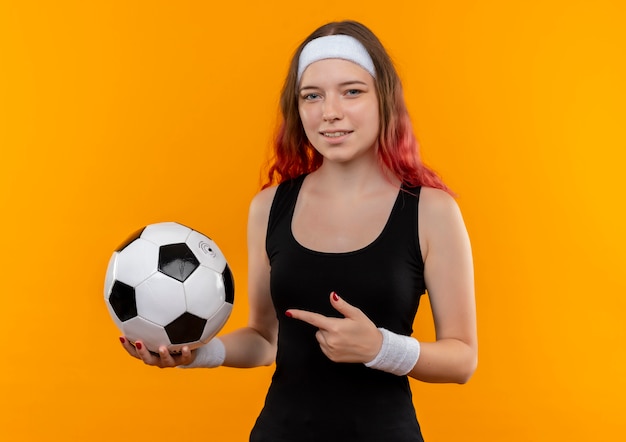 Mujer joven fitness en ropa deportiva sosteniendo un balón de fútbol apuntando con el dedo índice a él sonriendo de pie sobre la pared naranja
