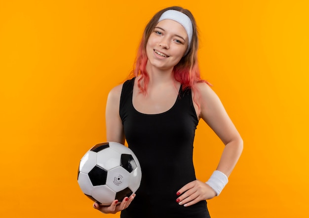 Mujer joven fitness en ropa deportiva sosteniendo un balón de fútbol con cara feliz sonriendo alegremente de pie sobre la pared naranja