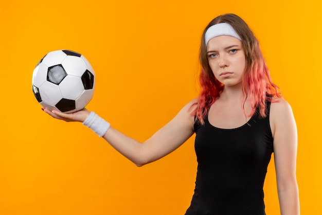 Foto gratuita mujer joven fitness en ropa deportiva sosteniendo un balón de fútbol en la mano con cara seria de pie sobre la pared naranja