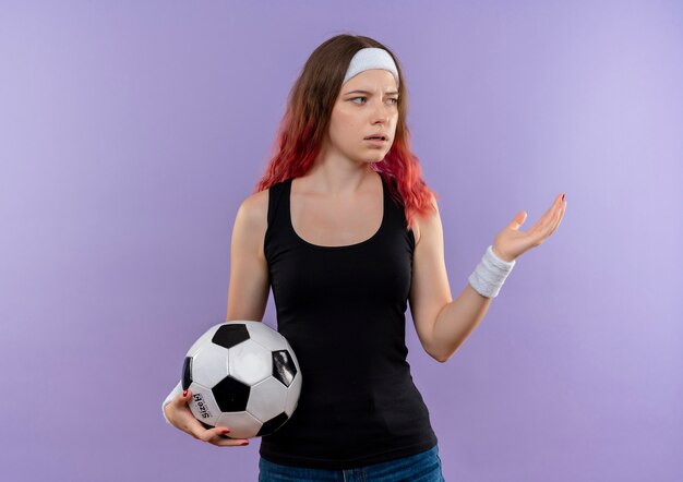 Mujer joven fitness en ropa deportiva sosteniendo un balón de fútbol mirando a un lado confundido parado sobre la pared púrpura