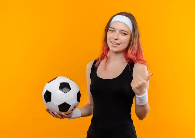 Mujer joven fitness en ropa deportiva sosteniendo un balón de fútbol sonriendo con cara feliz mostrando los pulgares para arriba sobre la pared naranja