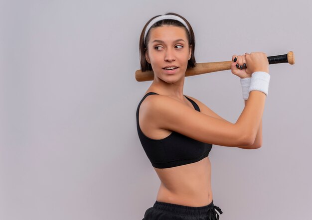 Mujer joven fitness en ropa deportiva sosteniendo un bate de béisbol mirando a un lado con expresión segura sonriendo de pie sobre la pared blanca