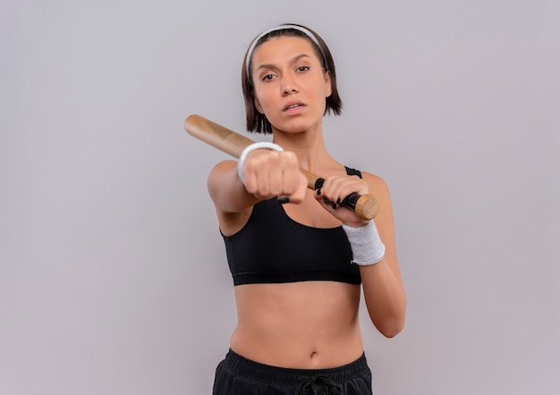 Mujer joven fitness en ropa deportiva sosteniendo un bate de béisbol mostrando su puño con cara seria de pie sobre la pared blanca