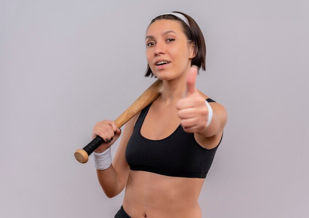 Mujer joven fitness en ropa deportiva sosteniendo un bate de béisbol sonriendo confiado mostrando los pulgares para arriba de pie sobre la pared blanca