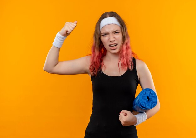 Mujer joven fitness en ropa deportiva sosteniendo estera de yoga levantando el puño emocionado y feliz de pie sobre la pared naranja