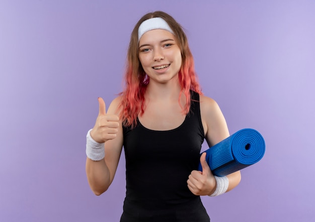 Mujer joven fitness en ropa deportiva sosteniendo estera de yoga sonriendo alegremente mostrando los pulgares para arriba de pie sobre la pared púrpura