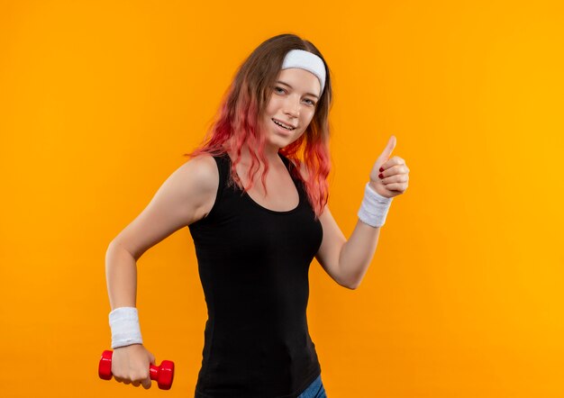 Mujer joven fitness en ropa deportiva sosteniendo mancuerna sonriendo alegremente mostrando los pulgares para arriba de pie sobre la pared naranja