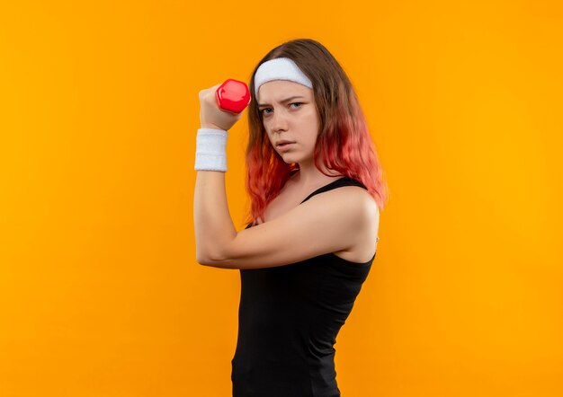 Mujer joven fitness en ropa deportiva sosteniendo mancuernas haciendo ejercicios de potencia con rostro serio parado sobre pared naranja