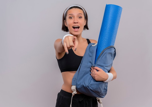 Mujer joven fitness en ropa deportiva sosteniendo mochila con estera de yoga feliz y sorprendido apuntando con índice a la cámara de pie sobre la pared blanca