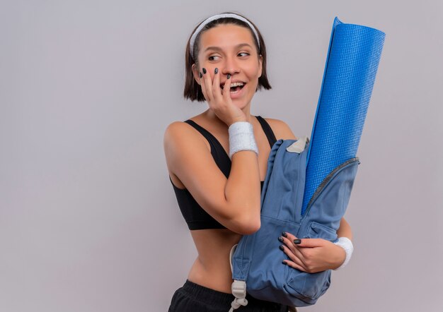 Mujer joven fitness en ropa deportiva sosteniendo la mochila con estera de yoga mirando a un lado sonriendo alegremente de pie sobre la pared blanca