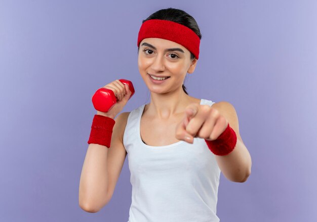 Mujer joven fitness en ropa deportiva sosteniendo pesas, apuntando con el dedo y sonriendo alegremente de pie sobre la pared púrpura