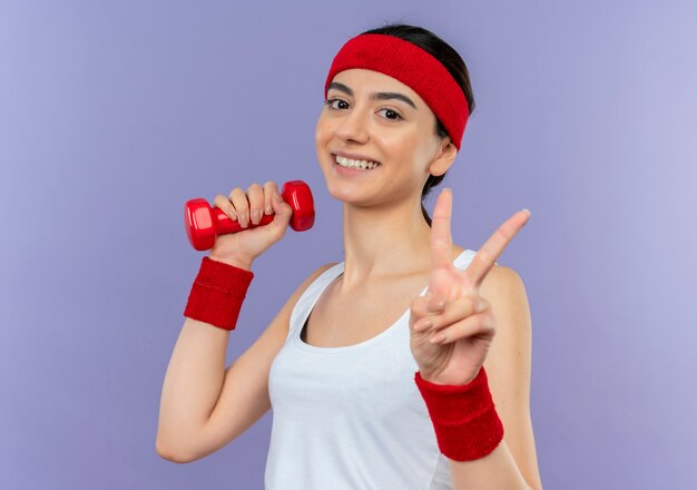 Mujer joven fitness en ropa deportiva sosteniendo pesas, posando y mostrando el signo de la victoria sonriendo alegremente de pie sobre la pared púrpura