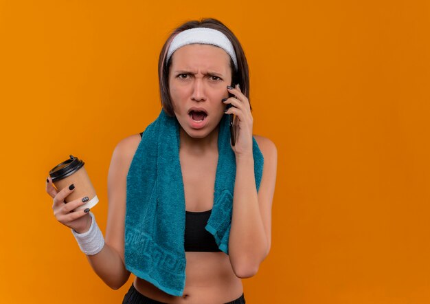 Mujer joven fitness en ropa deportiva con una toalla en el cuello sosteniendo la taza de café hablando por teléfono móvil con expresión agresiva de pie sobre la pared naranja
