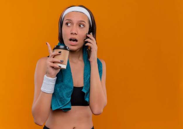 Mujer joven fitness en ropa deportiva con una toalla en el cuello sosteniendo la taza de café mirando confundido mientras habla por teléfono móvil de pie sobre la pared naranja