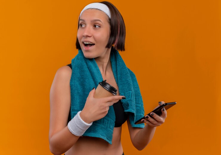 Foto gratuito mujer joven fitness en ropa deportiva con una toalla en el cuello sosteniendo una taza de café y un teléfono inteligente mirando a un lado sonriendo alegremente de pie sobre la pared naranja