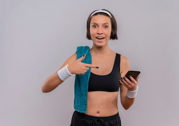 Mujer joven fitness en ropa deportiva con toalla en el hombro mostrando smartphone apuntando con el dedo sonriendo de pie sobre la pared blanca