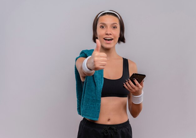 Mujer joven fitness en ropa deportiva con toalla en el hombro sosteniendo smartphone sonriendo mostrando los pulgares para arriba de pie sobre la pared blanca