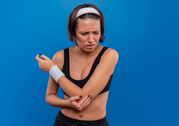 Mujer joven fitness en ropa deportiva tocando su codo con dolor de pie sobre la pared azul