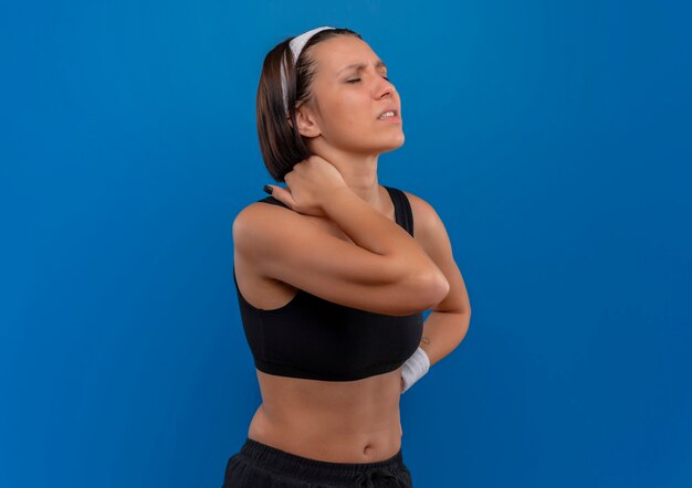 Mujer joven fitness en ropa deportiva tocando su cuello con dolor de pie sobre la pared azul