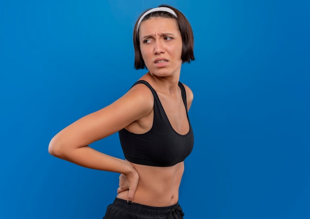 Mujer joven fitness en ropa deportiva tocando su espalda mirando a un lado confundido y muy ansioso de pie sobre la pared azul