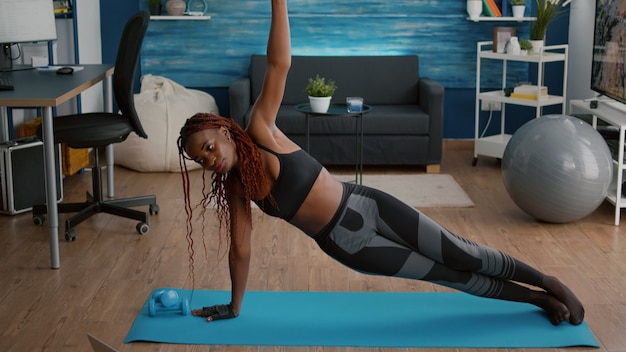 Mujer joven flexible calentamiento en mapa de yoga en la sala de estar de pie en la tabla lateral mirando video aeróbico en línea en la computadora portátil