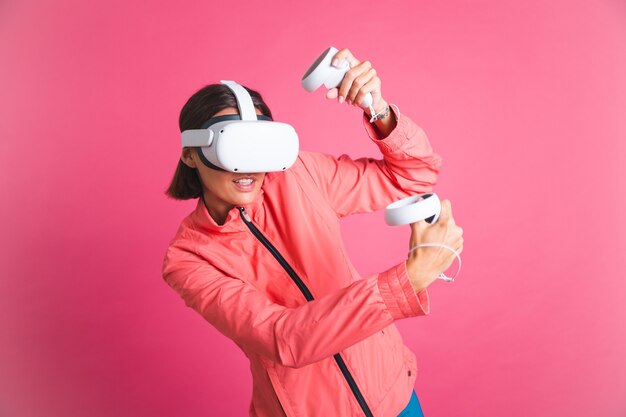 Mujer joven en forma con chaqueta deportiva y gafas de realidad virtual jugando juegos de lucha de boxeo en rosa
