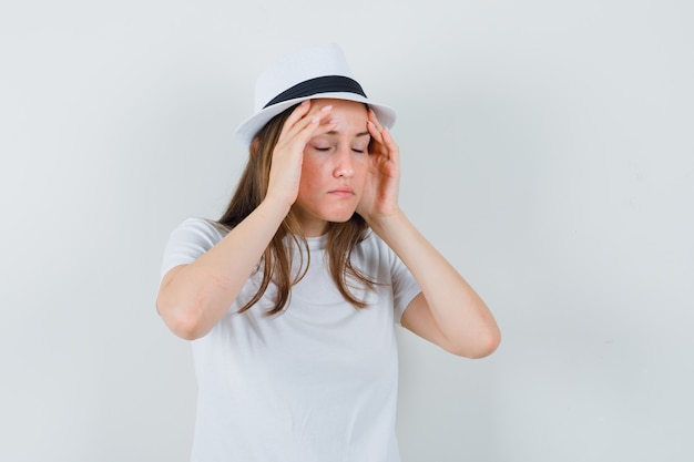 Mujer joven frotando las sienes en camiseta blanca, sombrero y aspecto cansado.