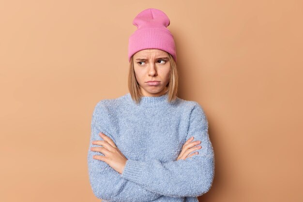 La mujer joven frustrada y molesta mantiene los brazos cruzados, se siente enojada u ofendida, frunce el ceño, se para en una postura cerrada, mira hacia otro lado, infelizmente, usa un sombrero rosa y un puente azul aislado sobre un fondo beige.