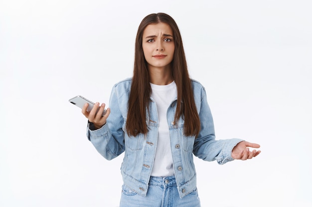 Mujer joven frustrada preocupada y disgustada con chaqueta de mezclilla, jeans, manos extendidas hacia los lados con consternación, sosteniendo el teléfono inteligente, frunciendo el ceño y sonriendo decepcionada, de pie fondo blanco confundido