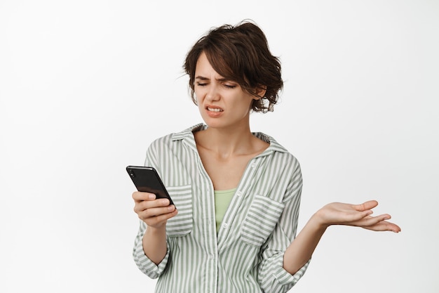 Mujer joven frustrada que mira la pantalla del teléfono móvil complicada, encogiéndose de hombros y frunciendo el ceño decepcionada, de pie en blanco.