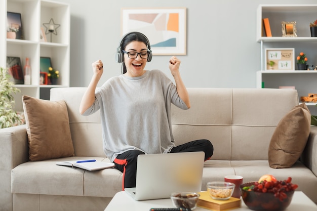 Mujer joven con gafas y auriculares sentado en el sofá detrás de la mesa de café mirando el portátil en la sala de estar