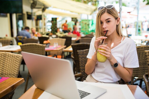 Mujer joven con gafas en la cabeza sonriendo con alegría, descansando en el café y navegando por internet usando una computadora portátil