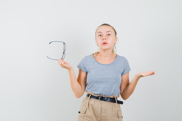 Mujer joven con gafas en camiseta, pantalón y mirada confundida. vista frontal.