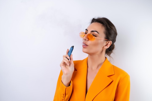 Foto gratuita una mujer joven con gafas elegantes de moda y una chaqueta de gran tamaño de color naranja brillante sobre un fondo blanco fuma un cigarrillo electrónico