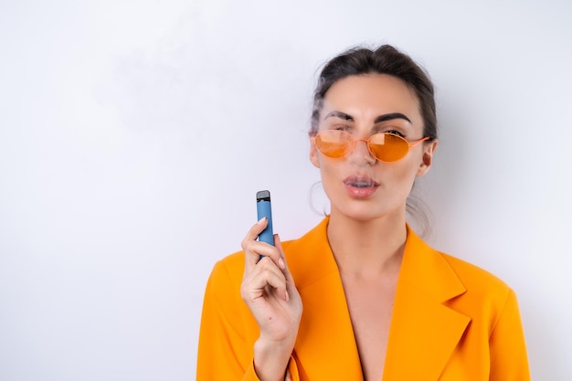 Una mujer joven con gafas elegantes de moda y una chaqueta de gran tamaño de color naranja brillante sobre un fondo blanco fuma un cigarrillo electrónico