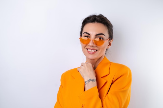 Mujer joven con gafas elegantes de moda y una chaqueta de gran tamaño de color naranja brillante sobre un fondo blanco