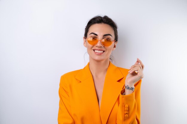 Mujer joven con gafas elegantes de moda y una chaqueta de gran tamaño de color naranja brillante sobre un fondo blanco