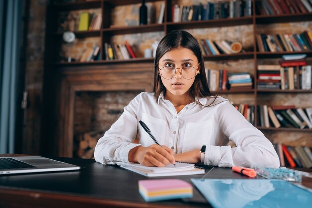 Mujer joven con gafas escribe en un cuaderno mientras está sentado en la mesa